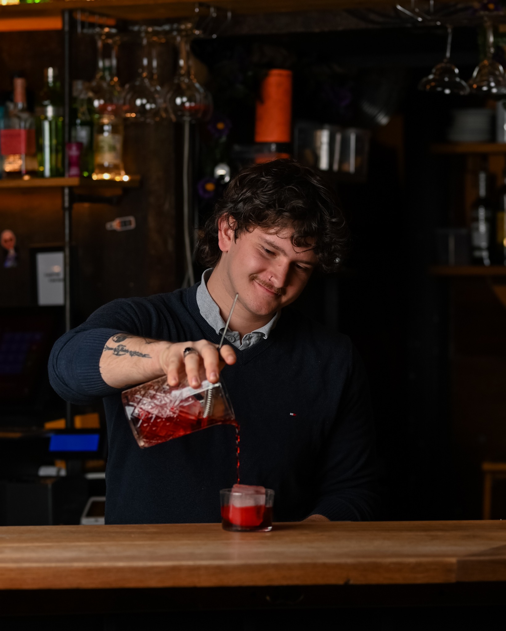 Barman z tatuażem nalewa czerwony koktajl z dzbanka do szklanki z lodem na drewnianym barze. W tle butelki i kieliszki.