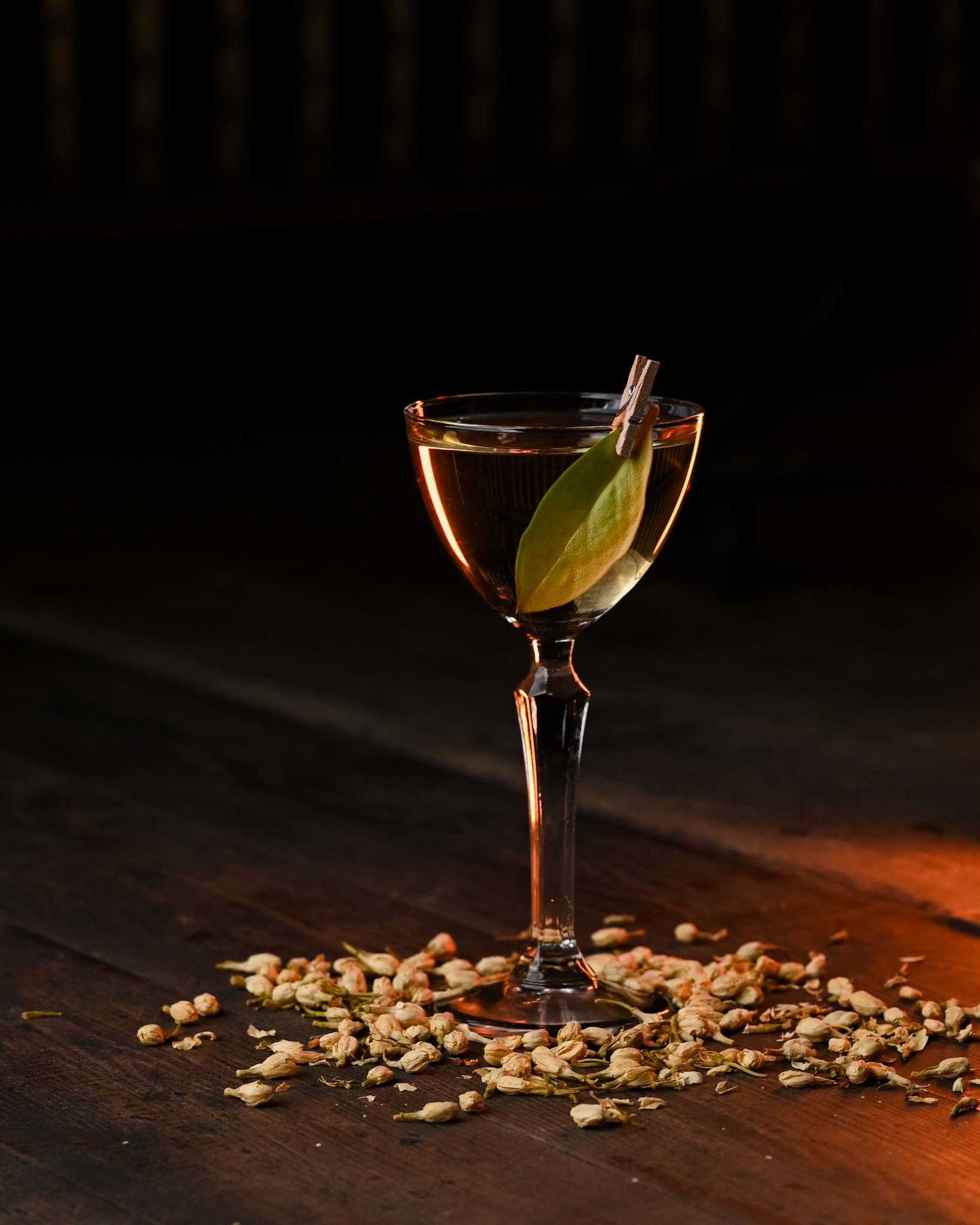 Elegancki koktajl w kieliszku z listkiem, ozdobiony klamerką, otoczony suszonymi kwiatami na drewnianym stole, w ciepłym, nastrojowym oświetleniu.