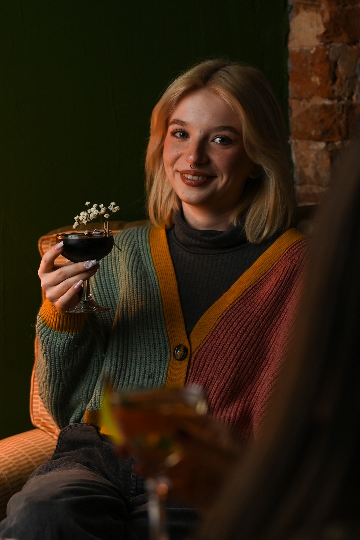 Uśmiechnięta blondynka z kolczykiem w nosie, trzyma koktajl z kwiatami. W tle cegła i zieleń. Klimatyczne oświetlenie, swobodna atmosfera.