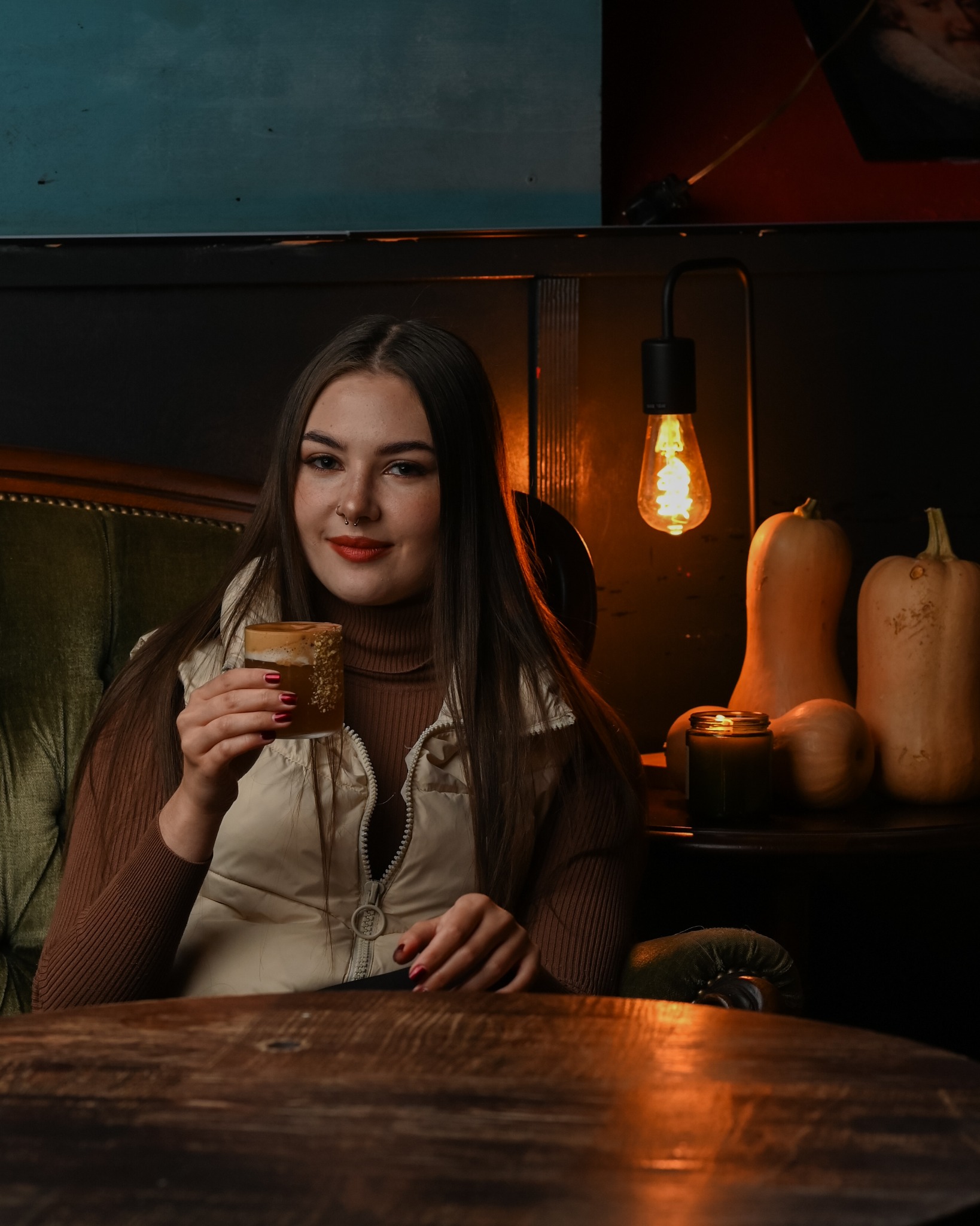Kobieta w beżowej kamizelce pije kawę przy drewnianym stole, w tle dekoracje z dyni i lampa edisona, tworząc przytulny klimat imprezy tematycznej.