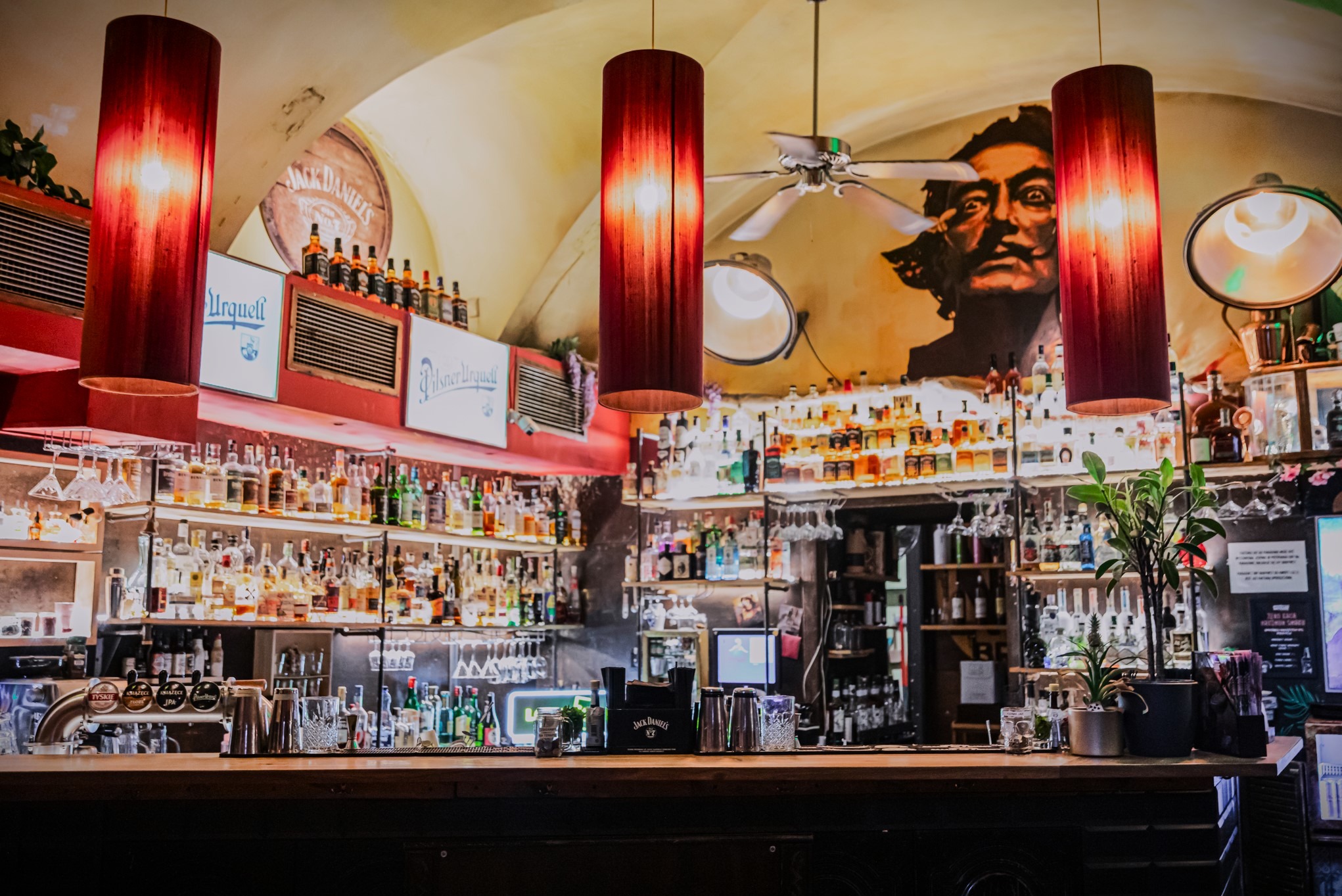 Wnętrze baru z bogato wyposażonym zapleczem, oświetlone czerwonymi lampami cylindrycznymi. Na ścianie mural z wizerunkiem Salvadora Dali.
