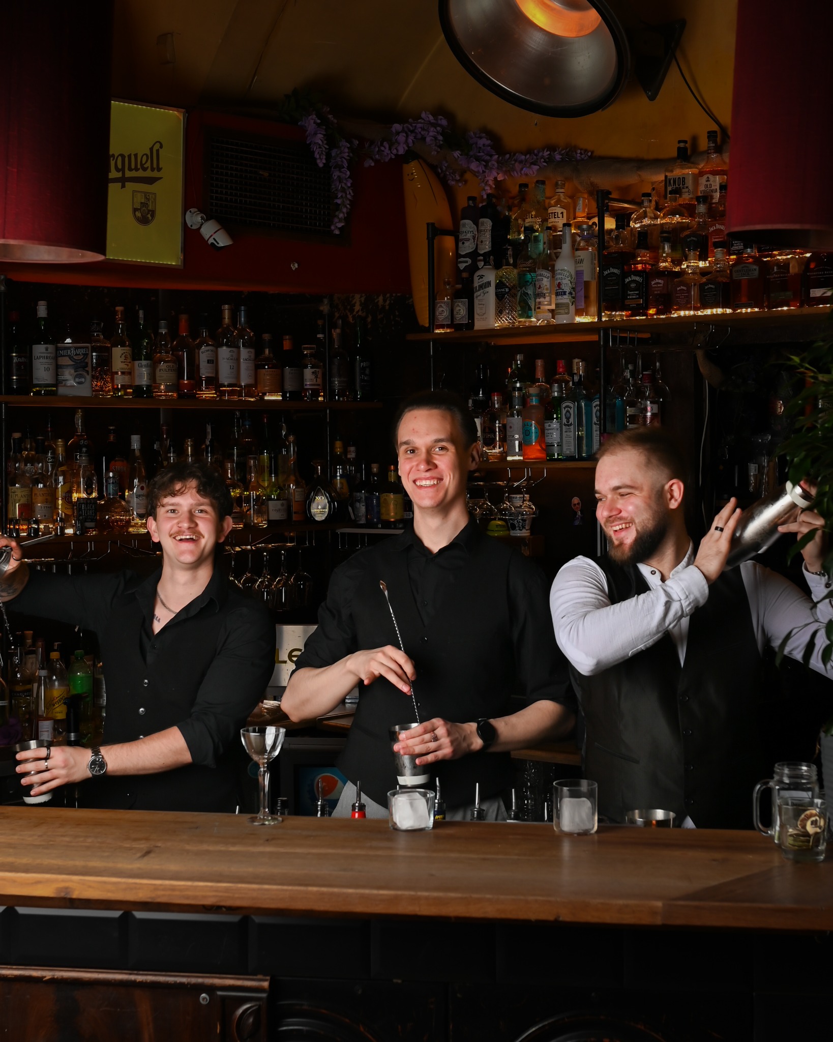 Uśmiechnięci barmani przygotowują drinki za drewnianym barem, w tle półki z butelkami alkoholi, oświetlenie punktowe. Klimatyczna atmosfera baru.