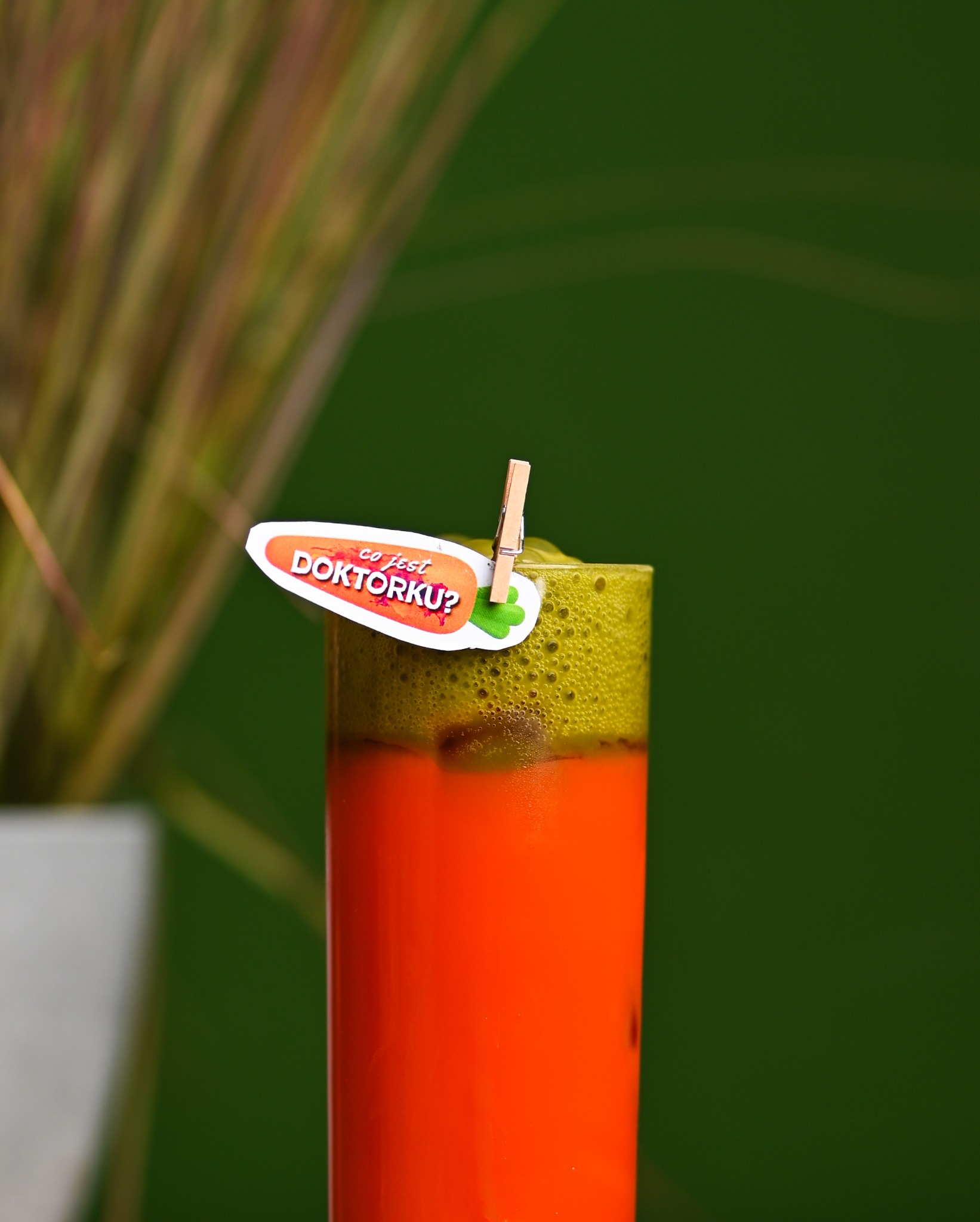 Pomarańczowo-zielony koktajl w szklance, ozdobiony etykietą 'Co jest doktorku?' przypiętą klamerką, na tle zielonej ściany i trawy w doniczce.