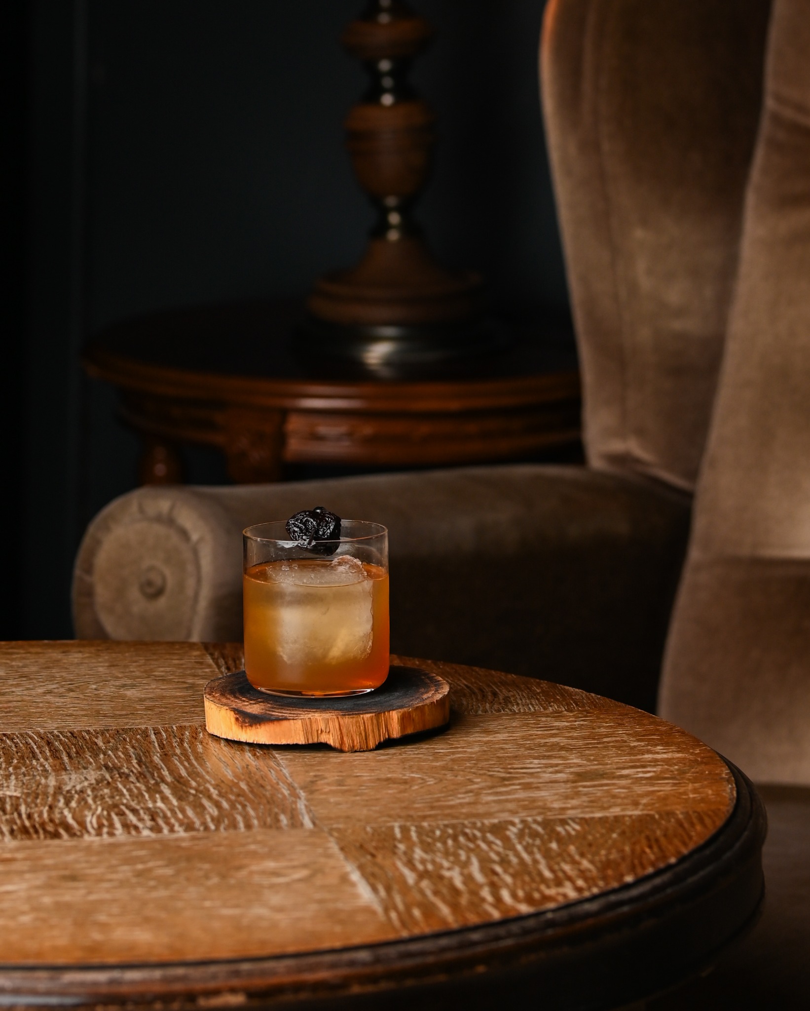 Kadr z drinkiem Old Fashioned z dużą kostką lodu i suszoną śliwką na drewnianym podkładce na stylowym stoliku. W tle fotel i lampa.