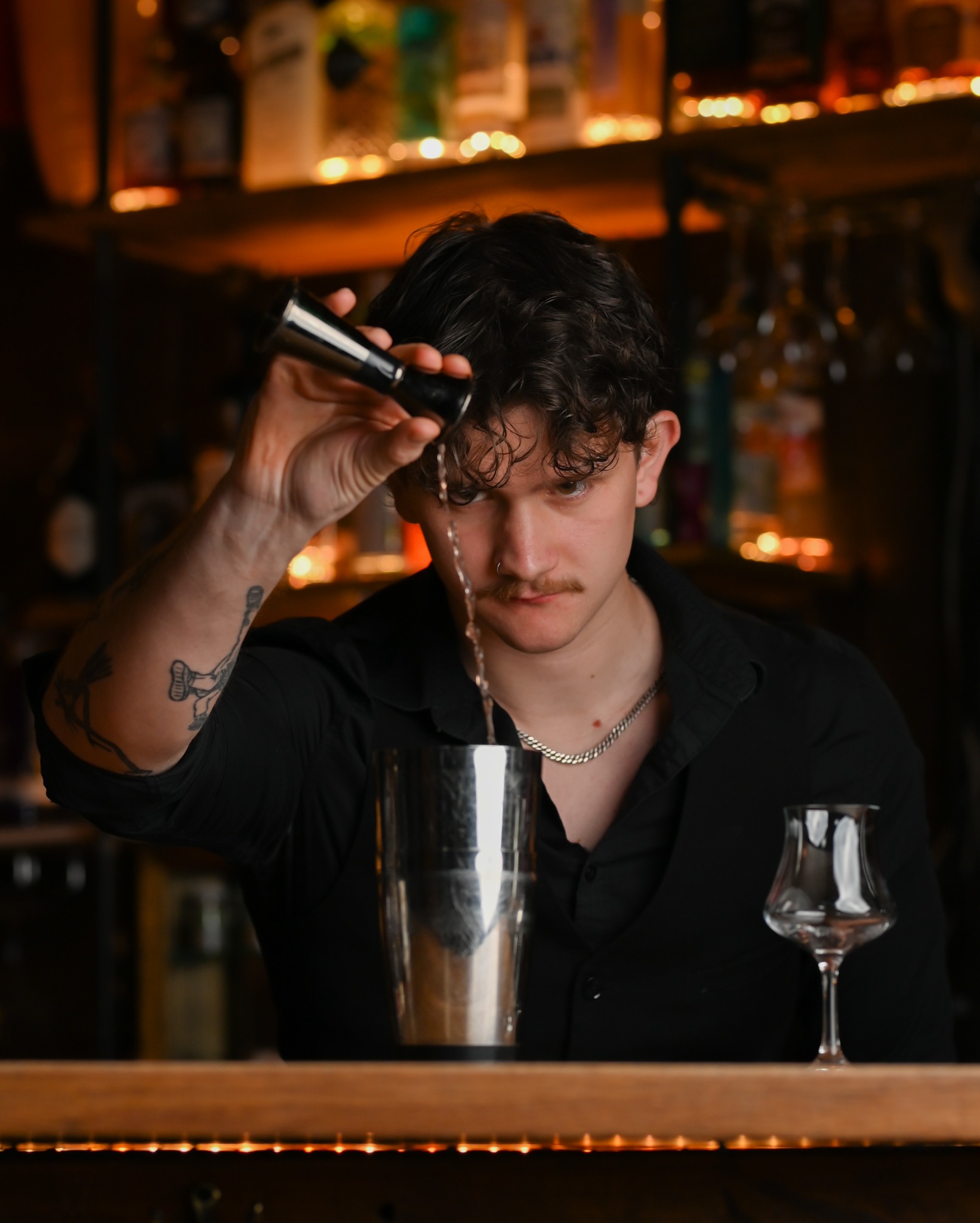 Barman w czarnej koszuli z tatuażem na ramieniu przygotowuje drinka, wlewając płyn z miarki do shakera na barze z podświetleniem LED.