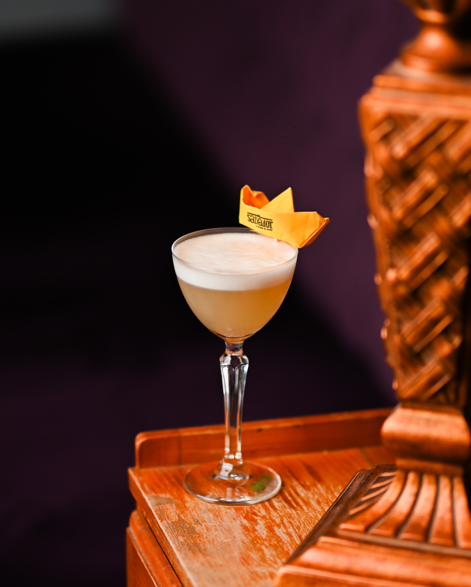 Kremowy koktajl w kieliszku z papierową łódką na drewnianym stoliku, w tle ciemna ściana. Dekoracja drinka na imprezę.