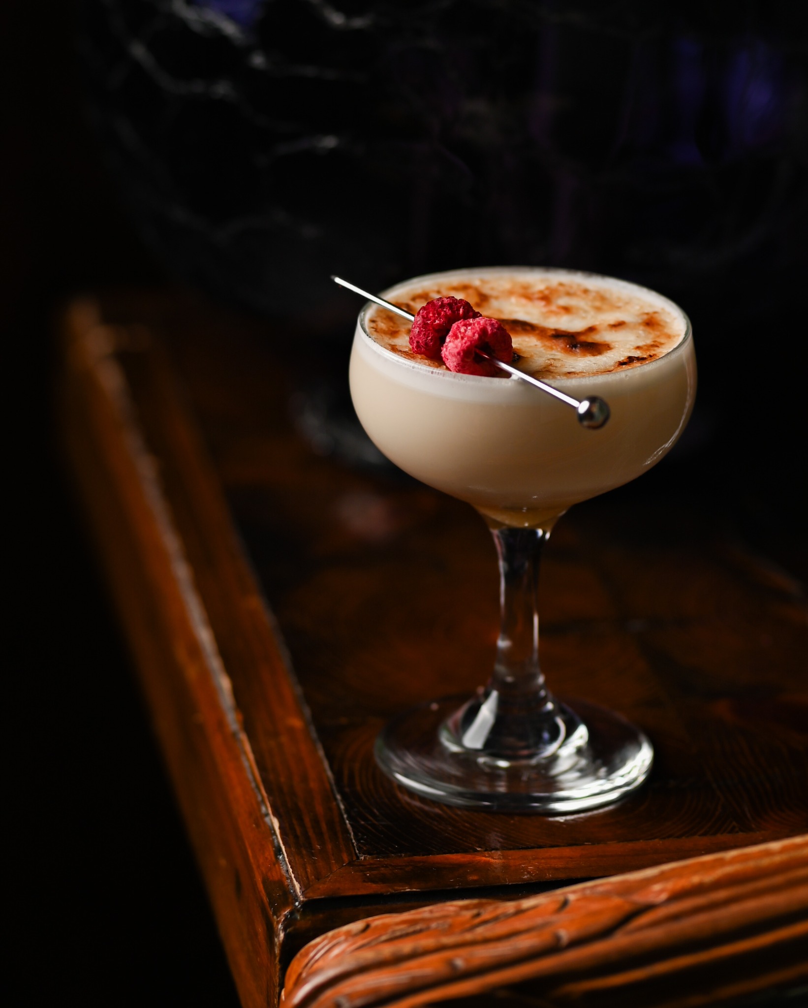 Kremowy koktajl w kieliszku z przypalonym wierzchem, udekorowany dwoma malinami na srebrnym szpikulcu, stoi na ciemnym, lakierowanym stoliku.