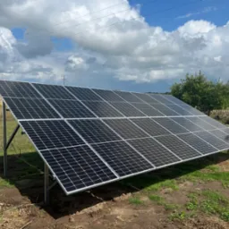 Panel słoneczny na metalowej konstrukcji, ustawiony na trawniku w wiejskim otoczeniu z widocznymi słupami energetycznymi w tle.