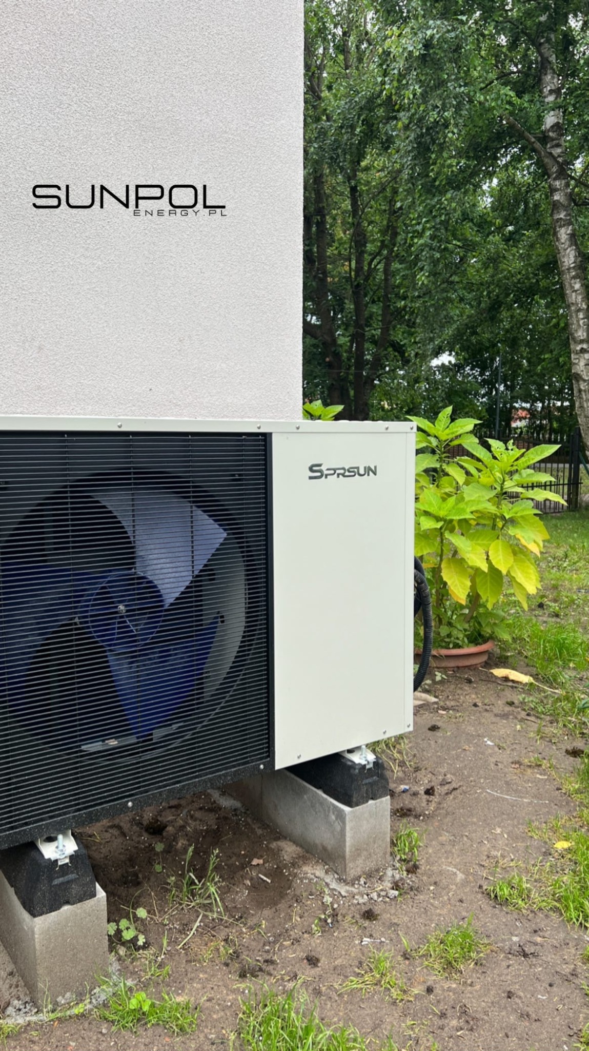 Zewnętrzna jednostka pompy ciepła Sprsun zainstalowana na betonowych podstawach, z widocznym wentylatorem i logo firmy Sunpol Energy na ścianie budynku w tle.