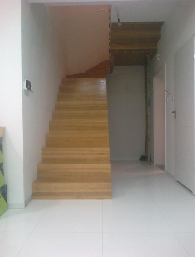 Proste, drewniane schody bez balustrady w jasnym wnętrzu z białymi płytkami podłogowymi, widoczne z dołu, z lekkim niedoświetleniem.