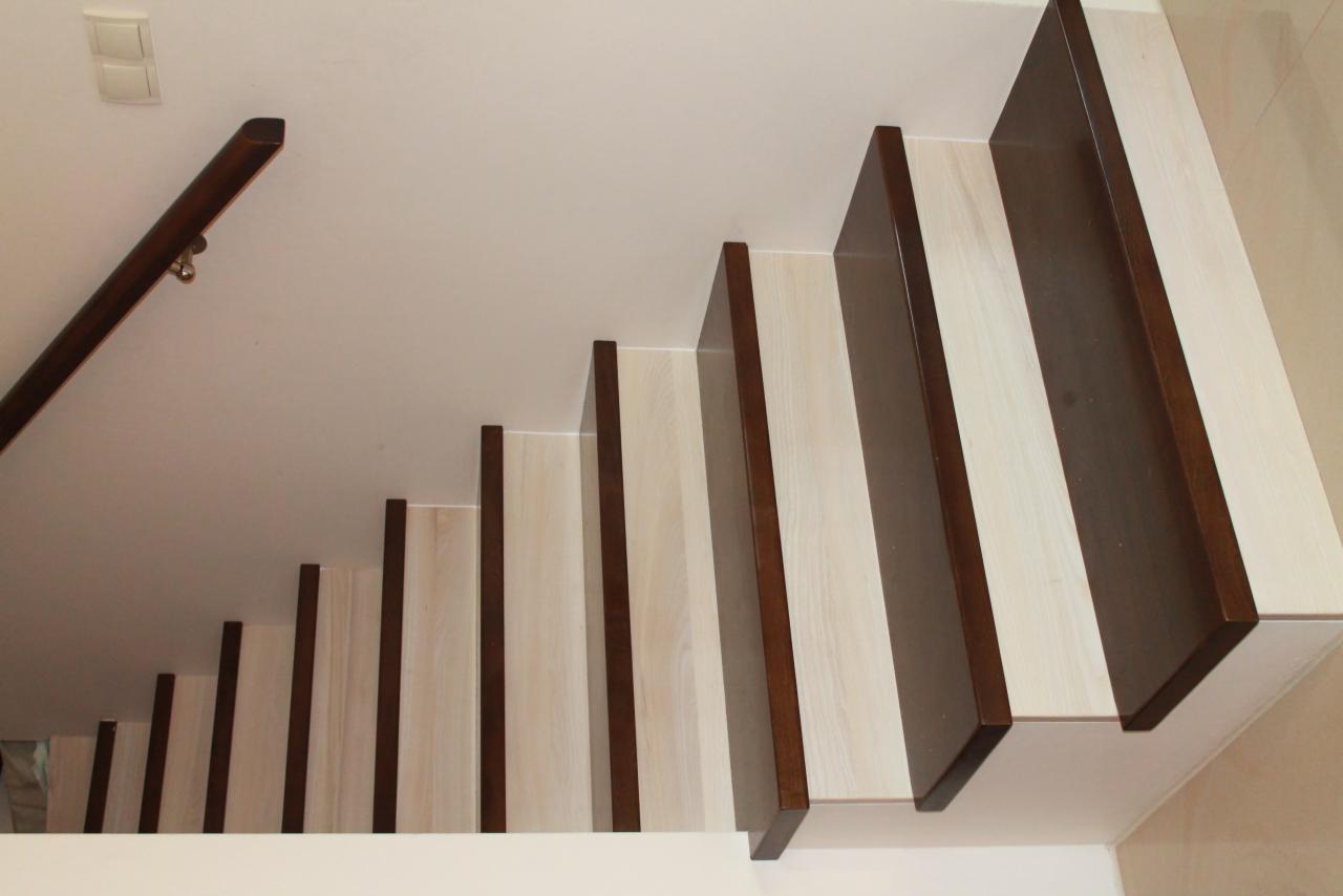 Drewniane schody z jasnymi stopniami i ciemnobrązowymi krawędziami, widok z góry, ukazujący poręcz po lewej stronie i biały sufit z oświetleniem.