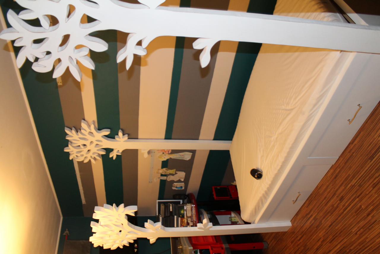 Białe łóżko dziecięce z ramą w kształcie drzewa, umieszczone na tle ściany w szerokie, pionowe pasy w odcieniach zieleni, szarości i bieli. Na ramie drzewa zawieszone dziecięce ubranka i zabawki.