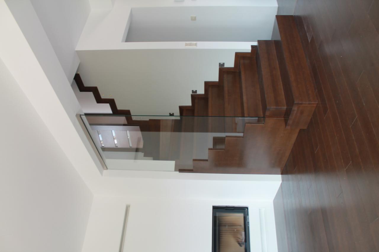 Nowoczesne schody drewniane z minimalistyczną szklaną balustradą w jasnym wnętrzu, widok z dołu, ukazujący detale wykończenia i oświetlenie.