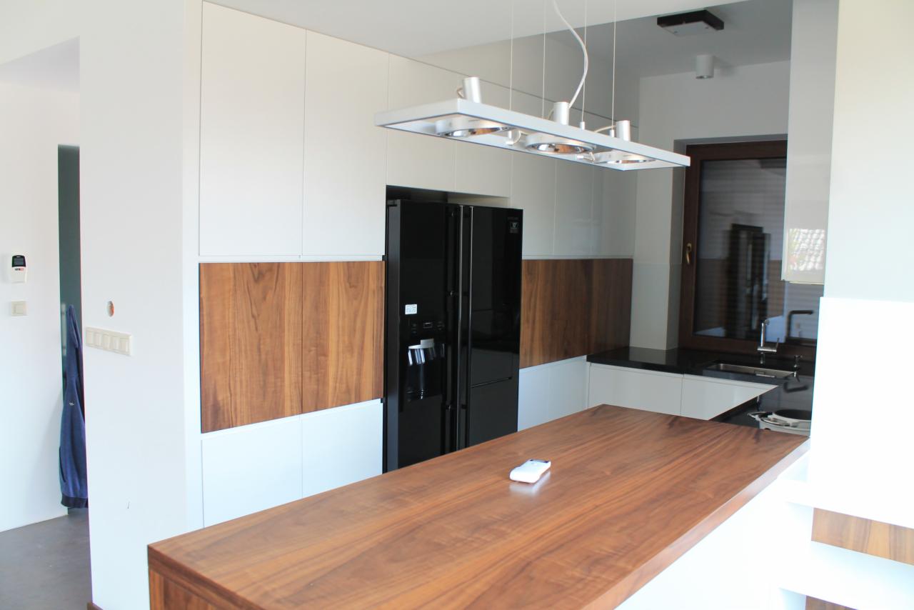 Nowoczesna kuchnia z białymi szafkami, drewnianymi akcentami i czarną lodówką, oświetlona wiszącą lampą. Widoczny drewniany blat wyspy kuchennej.