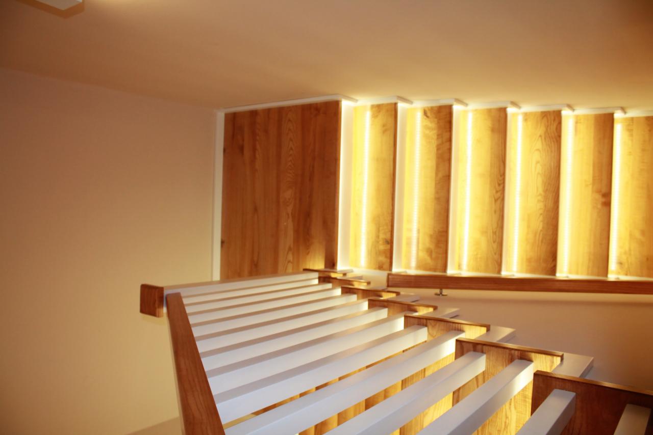 Nowoczesne schody z białymi stopniami i drewnianymi bokami, podświetlane pionowymi panelami z drewna i subtelnym oświetleniem LED.
