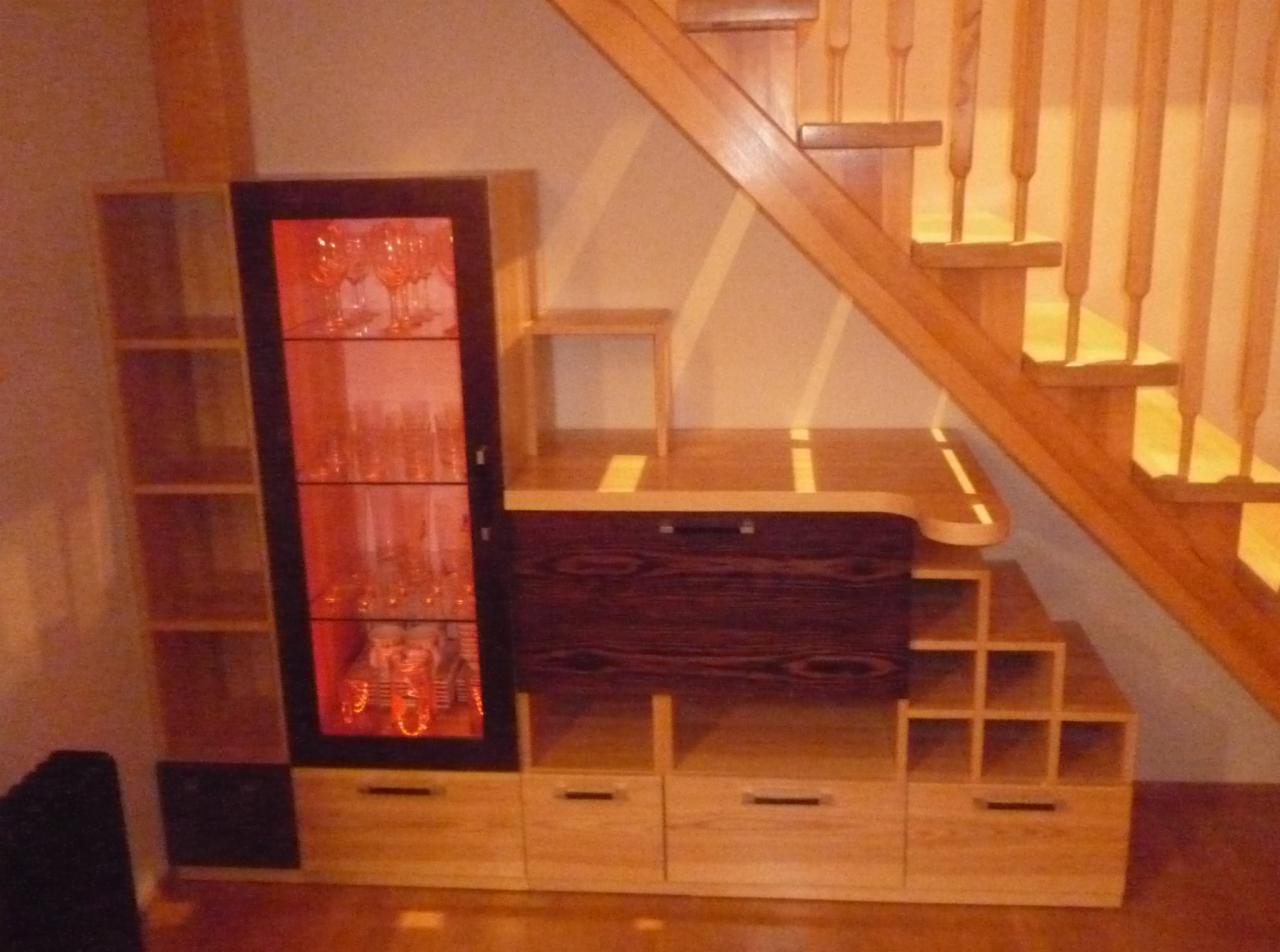 Zabudowa pod schodami z drewnianą szafką ekspozycyjną z podświetleniem, szufladami i półkami na wino, tworząca spójną całość z drewnianymi schodami.