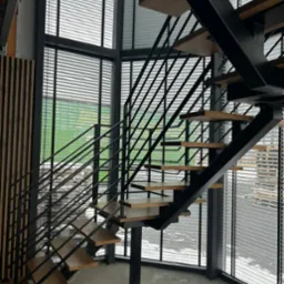 Nowoczesne schody z drewnianymi stopniami i czarną metalową konstrukcją, umieszczone w przeszklonej przestrzeni z widokiem na zewnątrz, oświetlone naturalnym światłem.