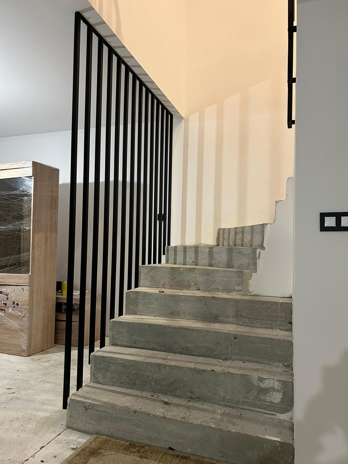 Surowe betonowe schody z czarną, pionową balustradą z metalowych prętów, widoczne ślady prac wykończeniowych na ścianie i meble w folii ochronnej.