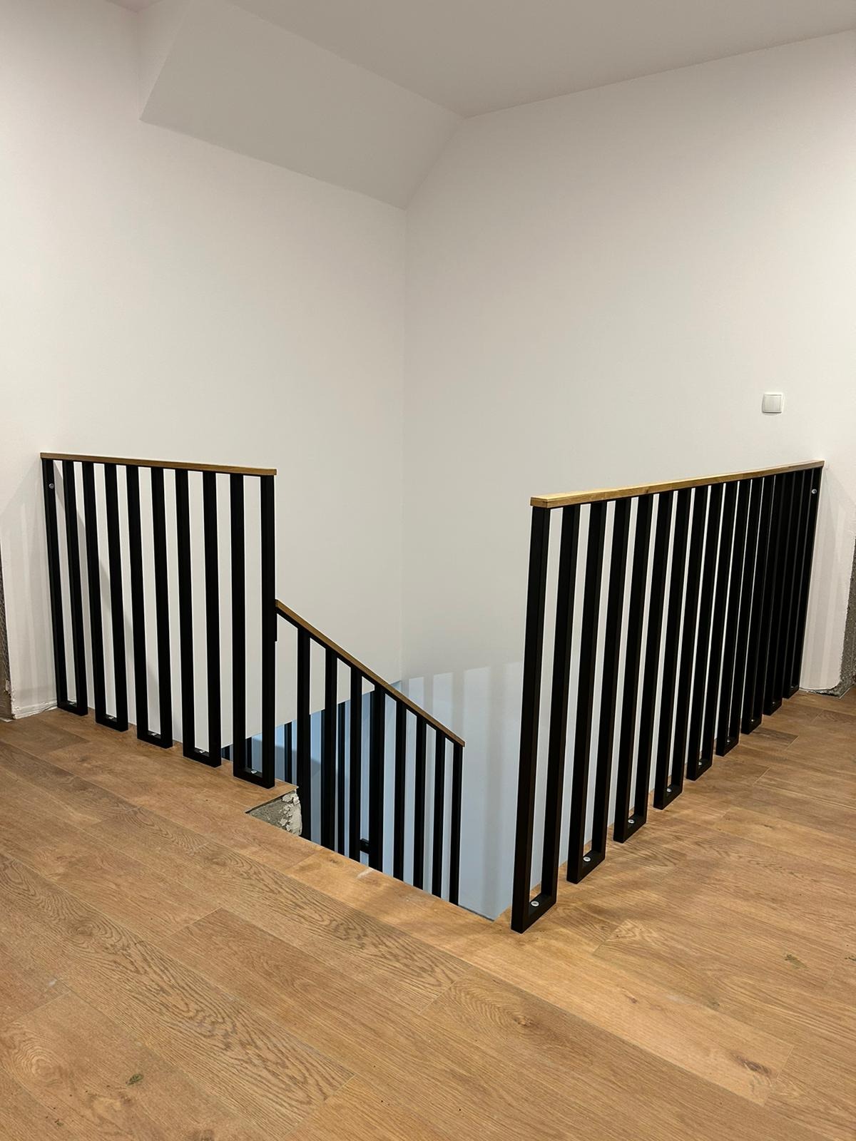 Nowoczesne schody z czarną metalową balustradą i drewnianym poręczą, widok z góry na drewnianą podłogę, białe ściany w tle.