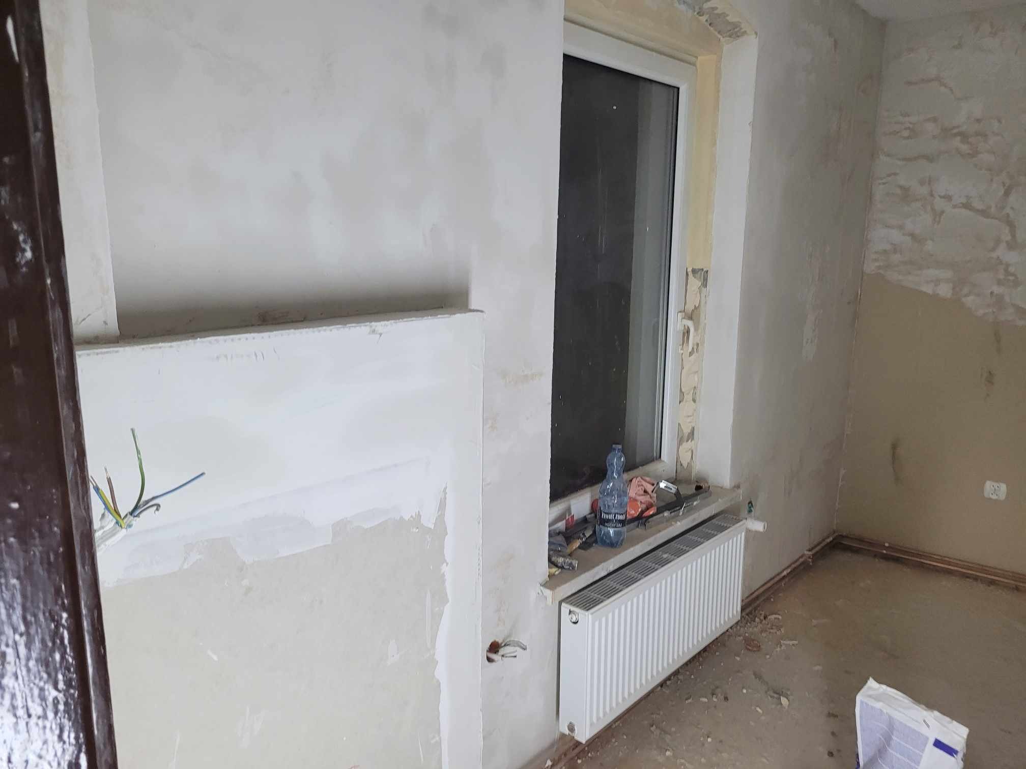Wnętrze pokoju w trakcie remontu: otynkowana ściana z wystającymi przewodami elektrycznymi, okno z grzejnikiem, butelka wody i narzędzia na parapecie, fragment ściany z resztkami starej farby.