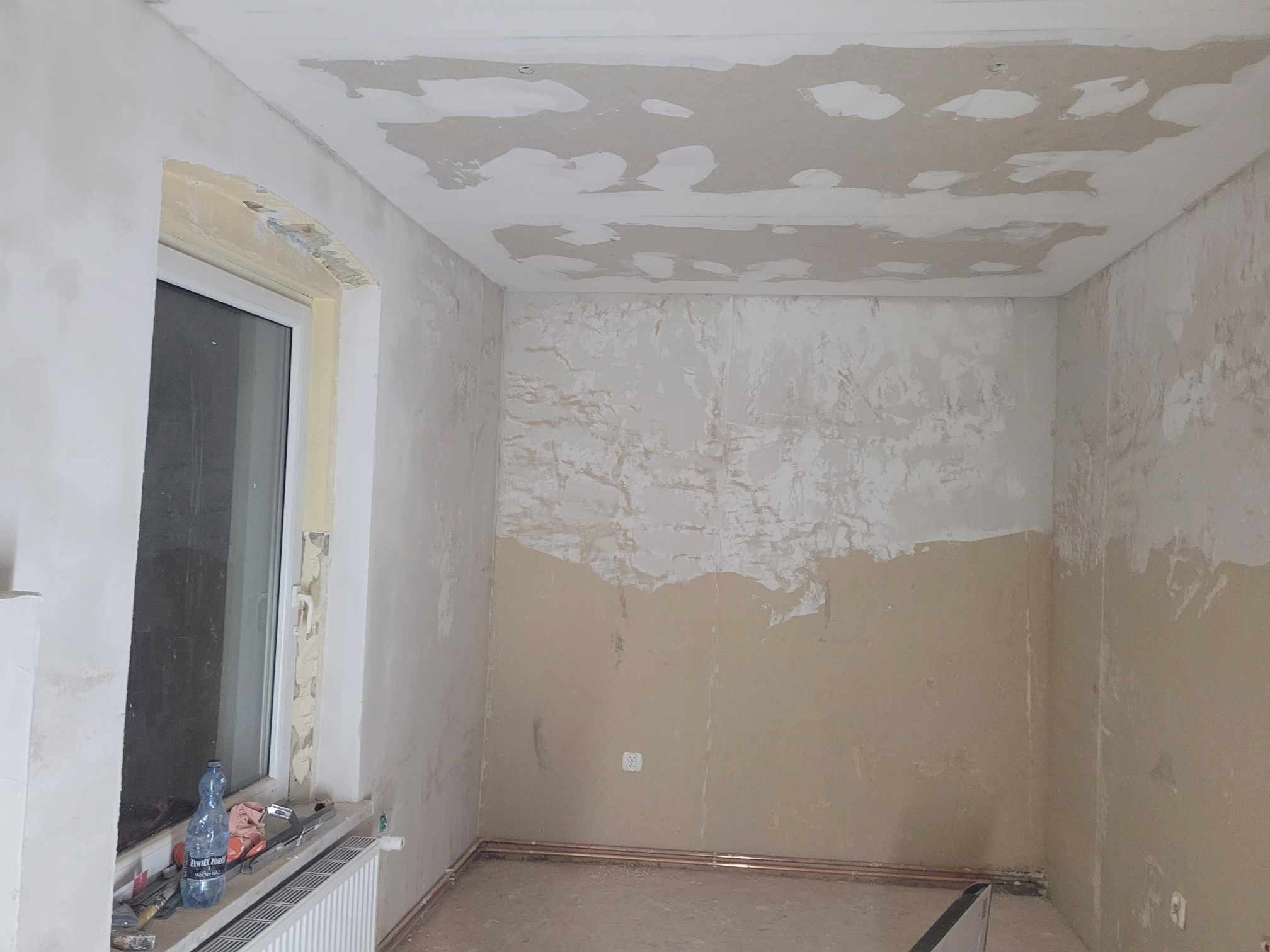 Pokój w trakcie remontu: ściany i sufit częściowo pokryte gładzią, widoczne narzędzia na parapecie okiennym, butelka wody mineralnej i grzejnik.