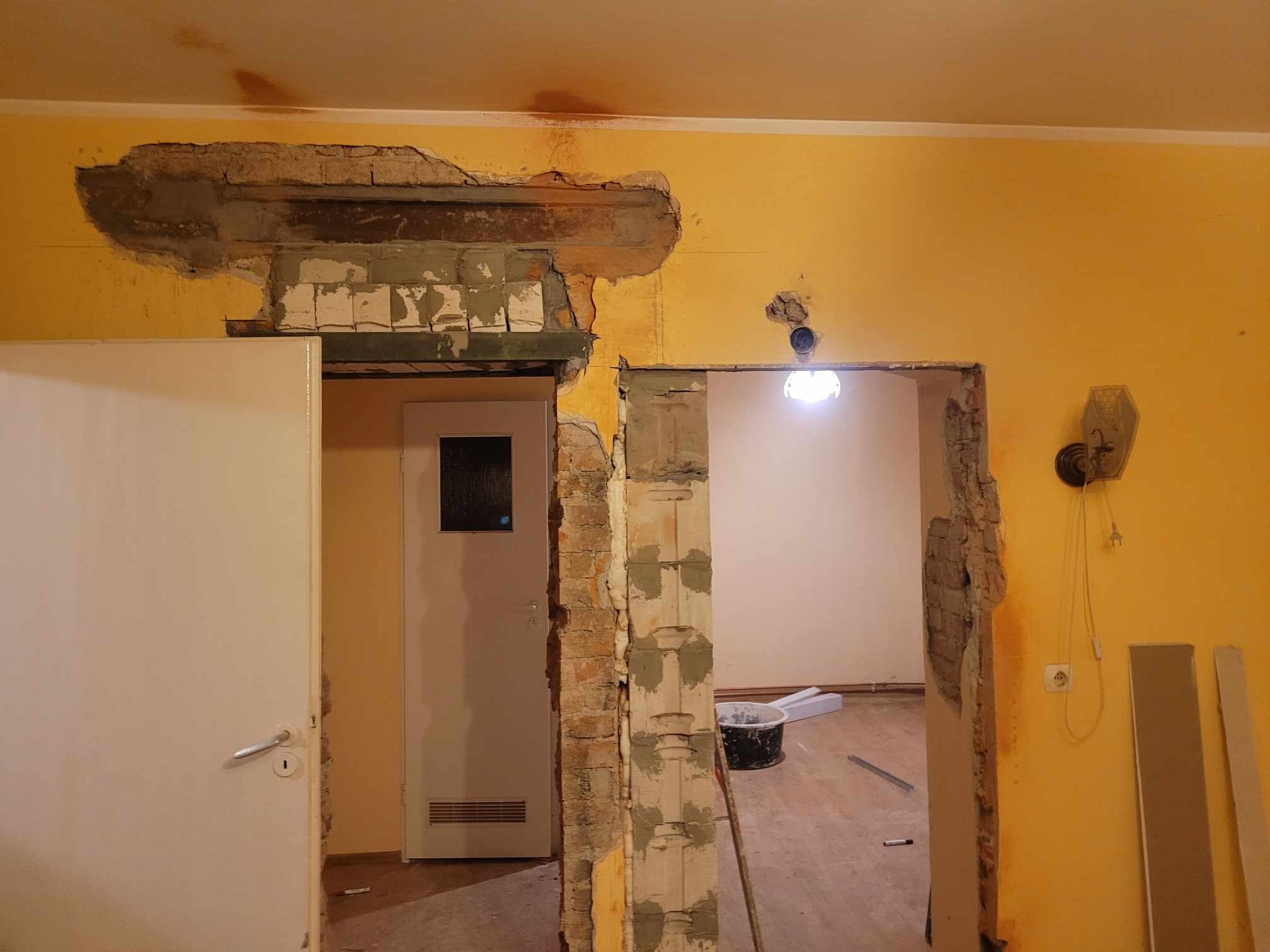 Prace remontowe: Wykucie otworu drzwiowego w żółtej ścianie, widoczne fragmenty cegieł i zaprawy, zarysowane nowe przejście.