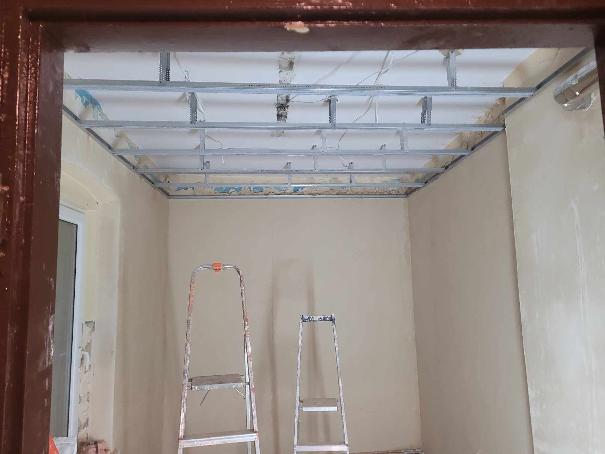 Stelaż sufitu podwieszanego z profili metalowych w trakcie montażu, widoczne dwie drabiny aluminiowe, ściany przygotowane do malowania.