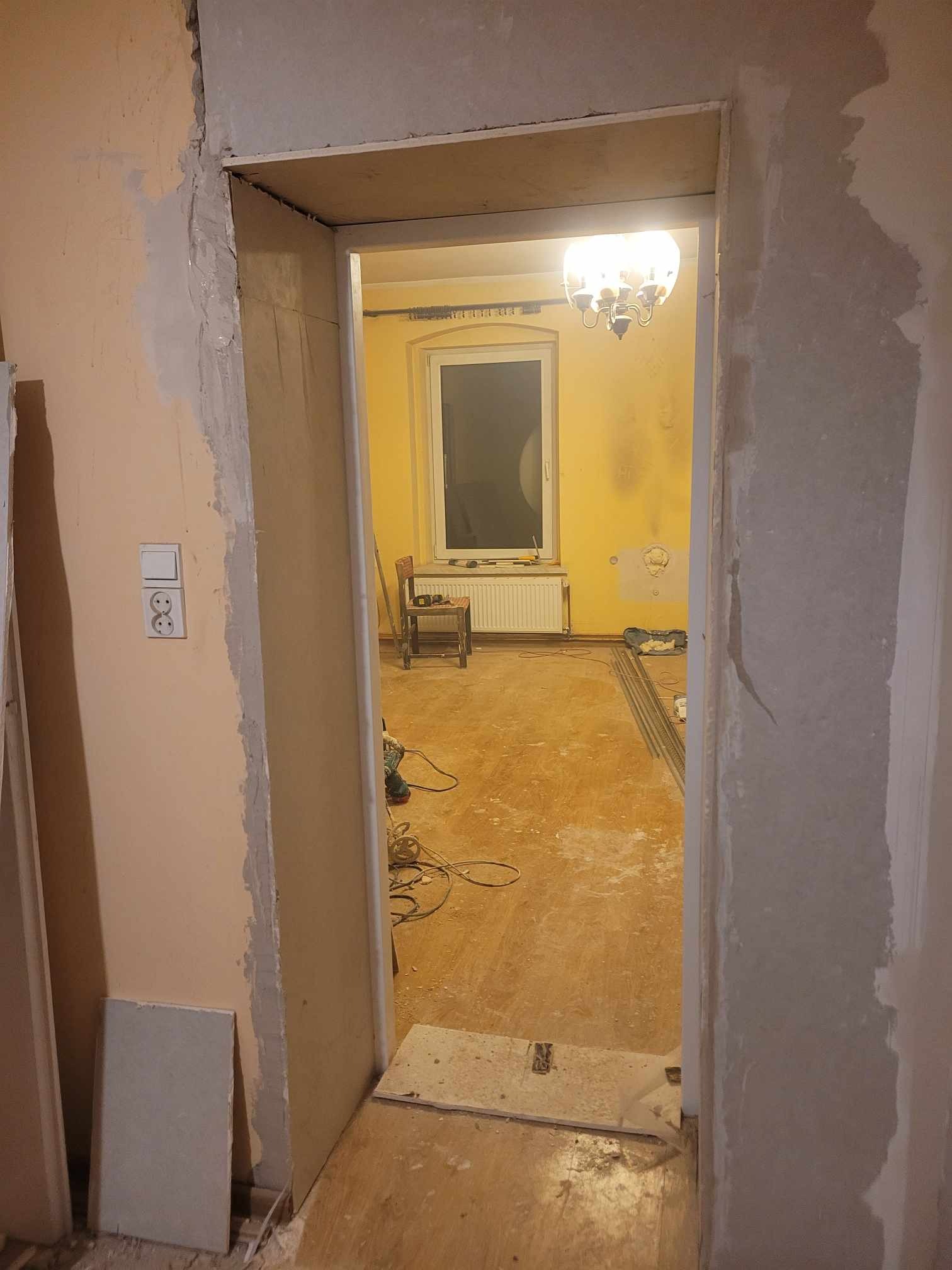 Widok przez futrynę drzwi na remontowany pokój z żółtymi ścianami, widoczne ślady po gładzi, listwy przypodłogowe i narzędzia.