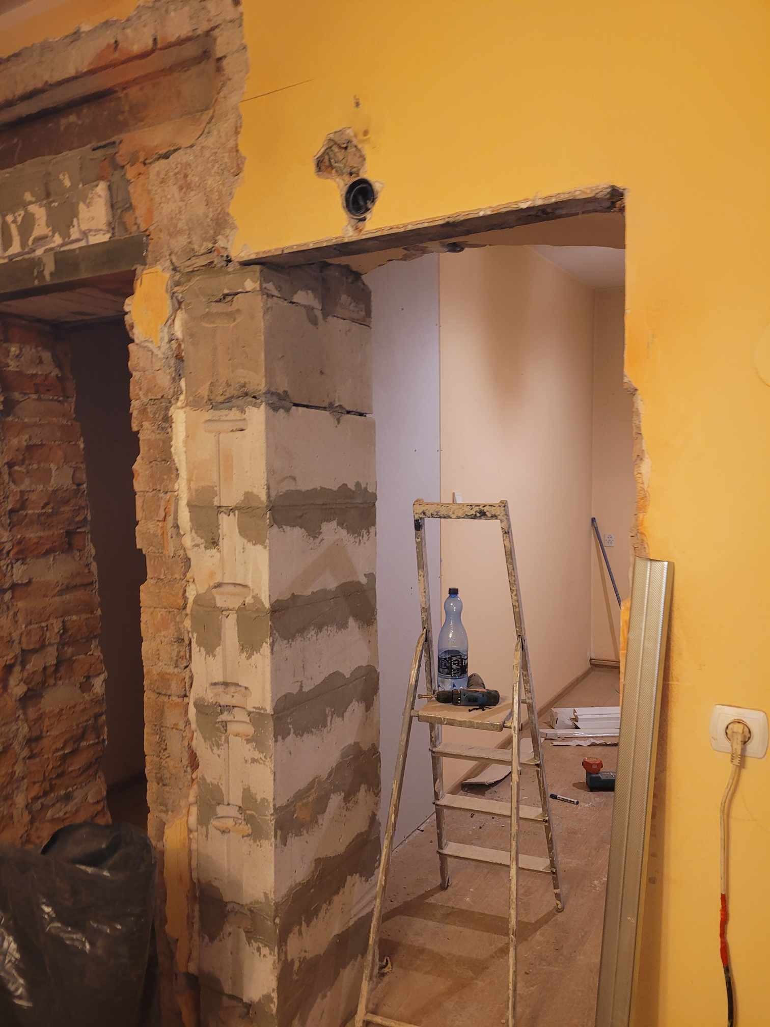 Wnętrze w trakcie remontu: odsłonięta cegła obok otynkowanego filaru, drabina z butelką wody i wiertarką, żółta ściana z wystającym gniazdkiem elektrycznym.