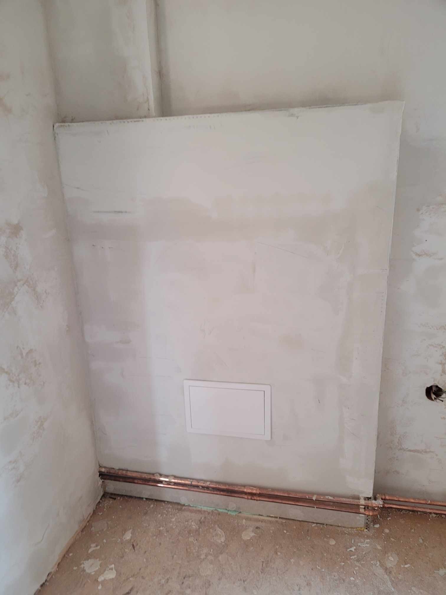 Nierówna, surowa ściana z gładzią gipsową przed malowaniem, z widocznymi miedzianymi rurami i małą, kwadratową klapą rewizyjną w dolnej części.