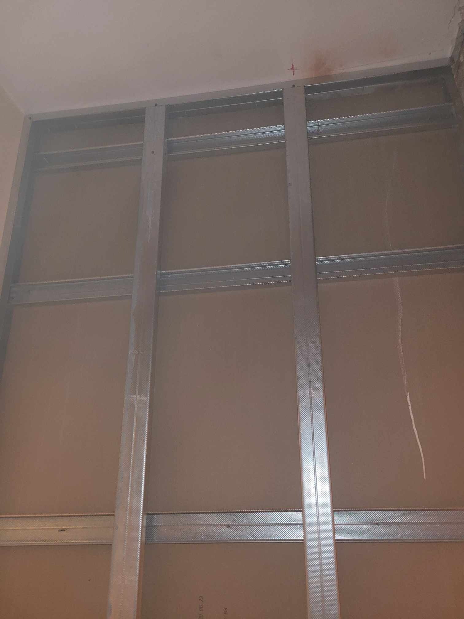 Konstrukcja metalowa pod płyty gipsowo-kartonowe na ścianie, widoczne profile pionowe i poziome, czerwony znak na suficie.