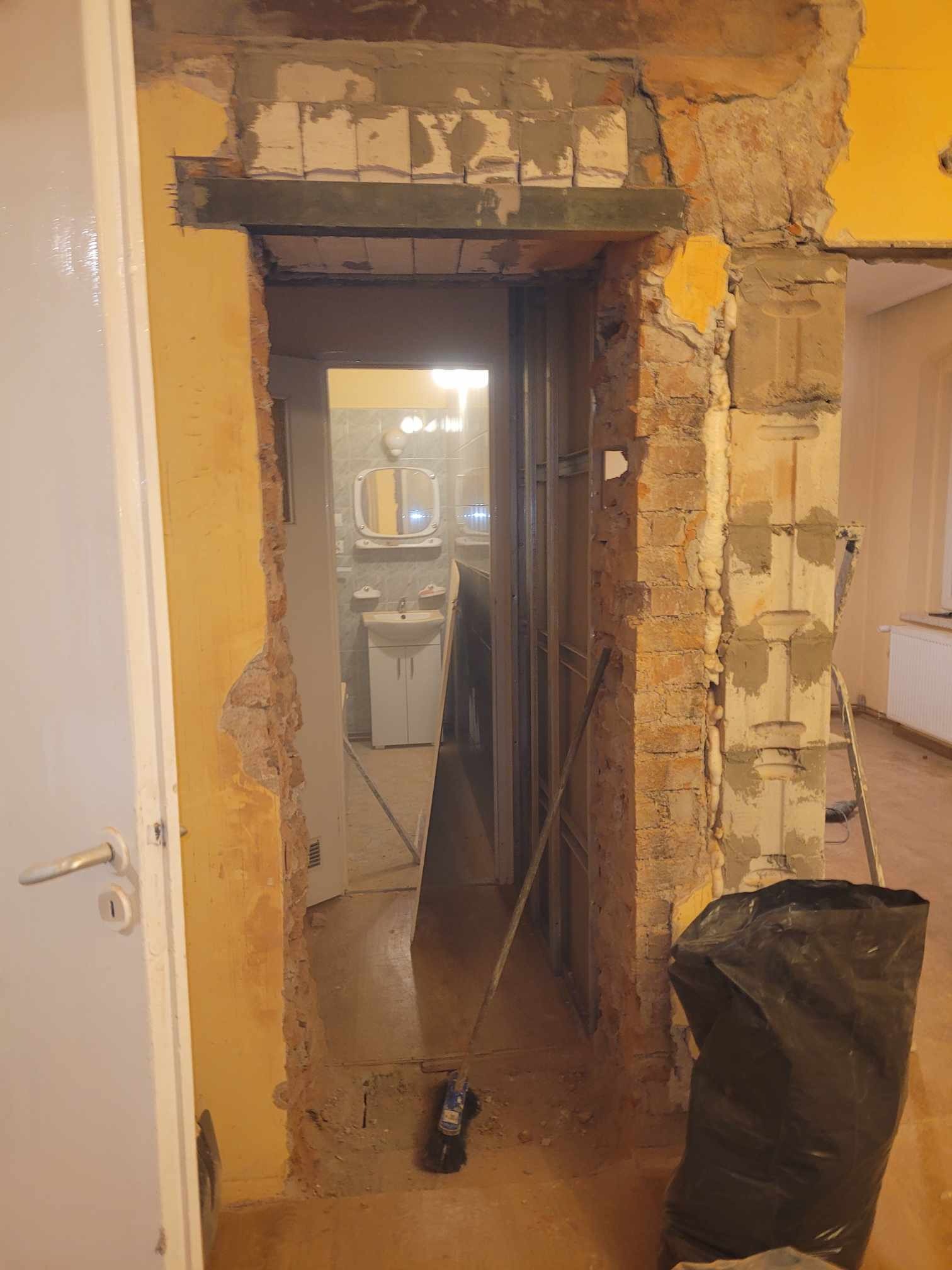 Otwór drzwiowy w trakcie remontu: odsłonięta cegła, fragmenty tynku, metalowa belka wzmacniająca nad otworem, w tle widoczna łazienka z białą umywalką i lustrem, na podłodze gruz i czarny worek...