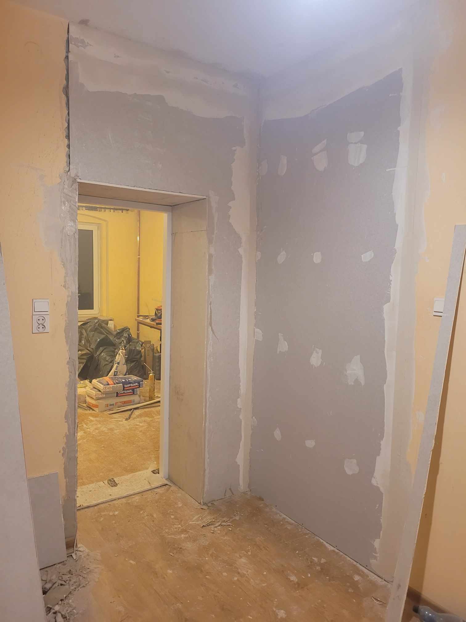 Wnętrze pokoju w trakcie remontu: ściany z nałożoną gładzią gipsową, widoczne worki z gipsem w przejściu do kolejnego pomieszczenia, przygotowane do dalszych prac wykończeniowych.