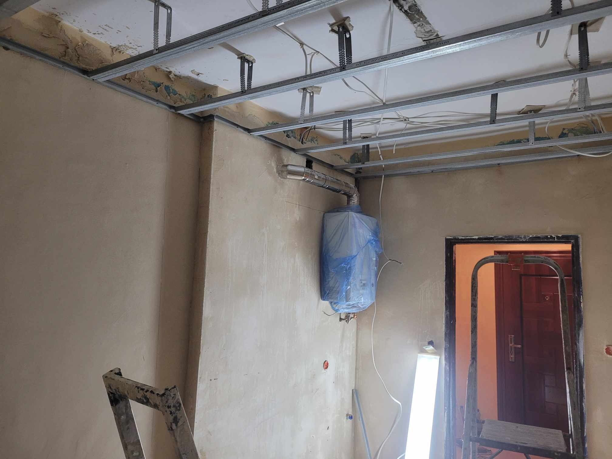 Wnętrze w trakcie remontu: surowy sufit podwieszany z metalowym stelażem, ściany w stanie surowym, zabezpieczony folią bojler z podłączonymi rurami, drabina malarska i prowizoryczne oświetlenie.