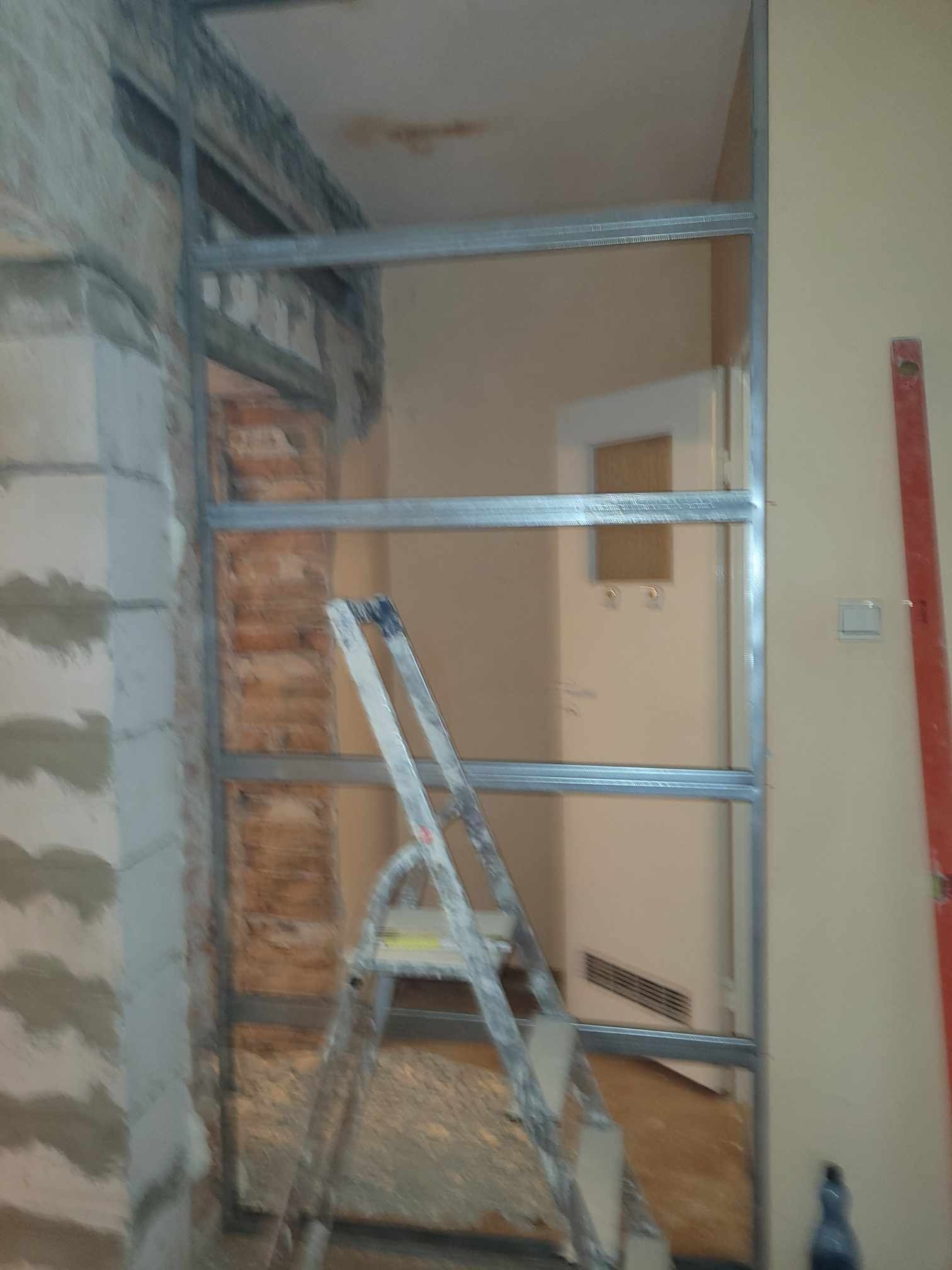 Metalowa konstrukcja szkieletowa w trakcie montażu, widoczny fragment ceglanej ściany i drabina, pomieszczenie w trakcie remontu.