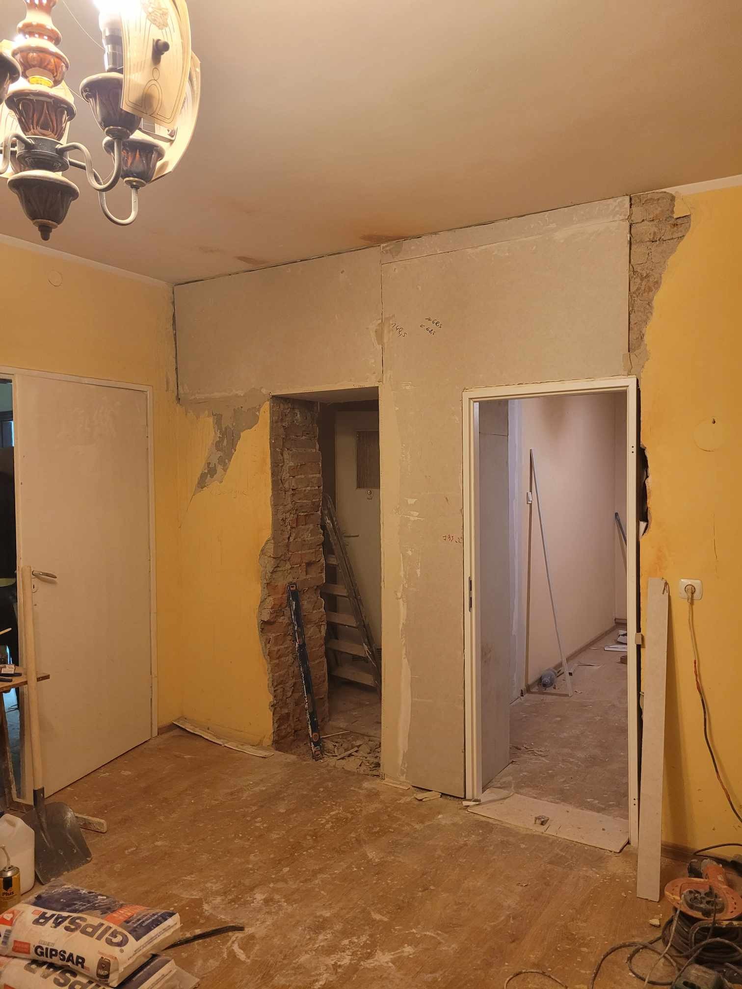 Wnętrze w trakcie remontu: odsłonięta cegła przy przejściu, częściowo położona gładź, worki z gipsem i narzędzia na podłodze, widoczna drabina w głębi pomieszczenia.