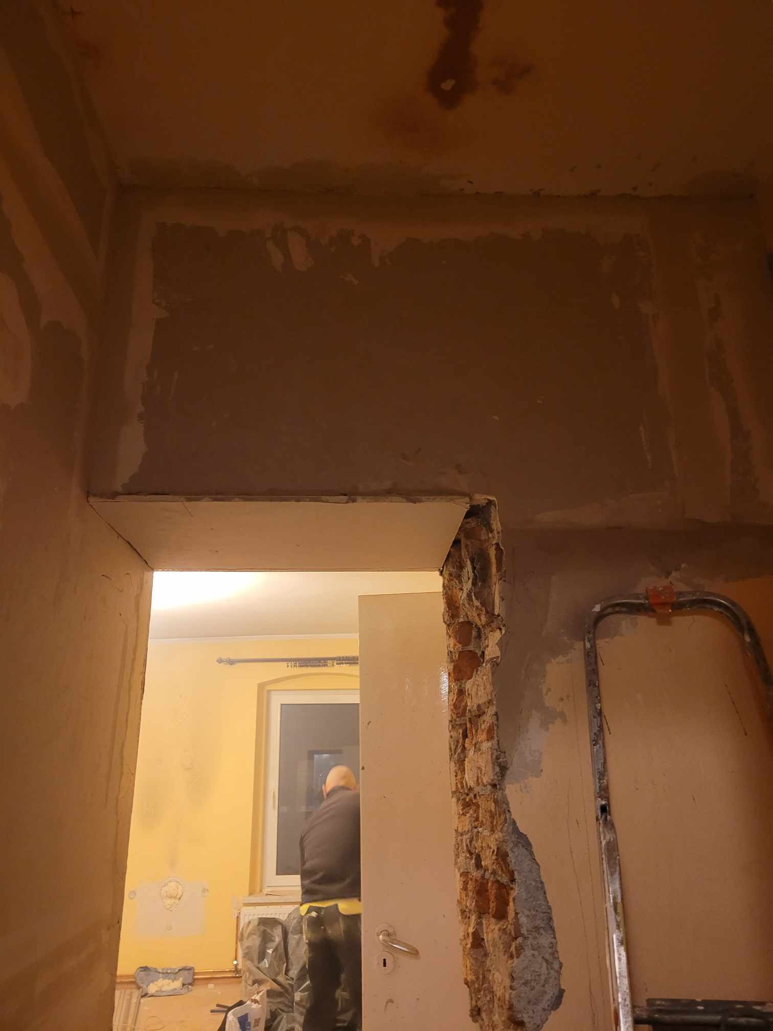 Fragment ściany z uszkodzonym tynkiem, widoczne cegły, częściowo zatynkowana ściana, drabina oparta o ścianę, widok na pokój z żółtymi ścianami i oknem.