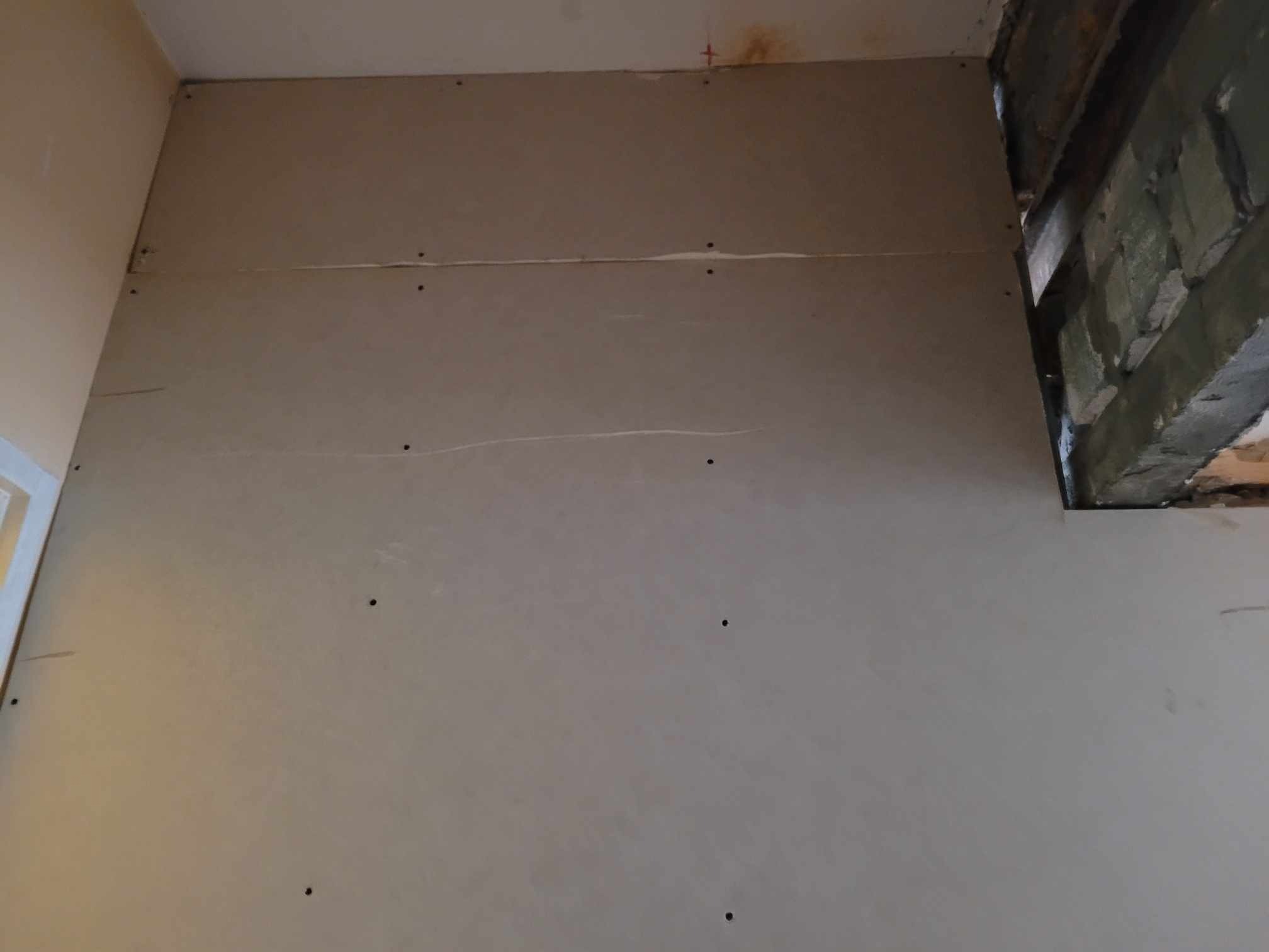 Płyta gipsowo-kartonowa zamontowana na ścianie z widocznymi śladami po wkrętach i niewielkim otworem odsłaniającym fragment muru z cegieł.