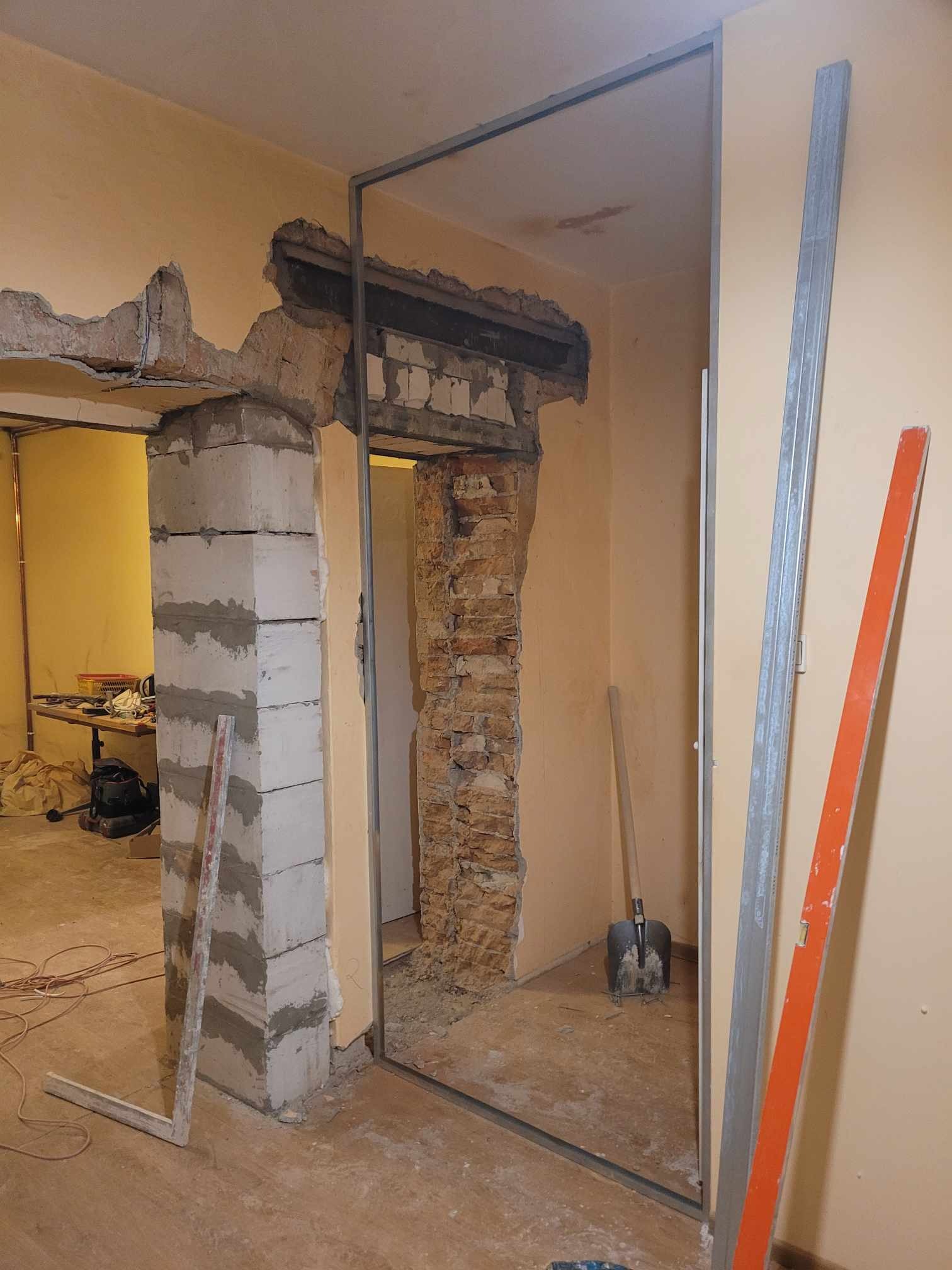 Przygotowanie otworu drzwiowego: usunięte fragmenty ściany, widoczne cegły i pustaki, metalowa rama ościeżnicy, narzędzia w tle.