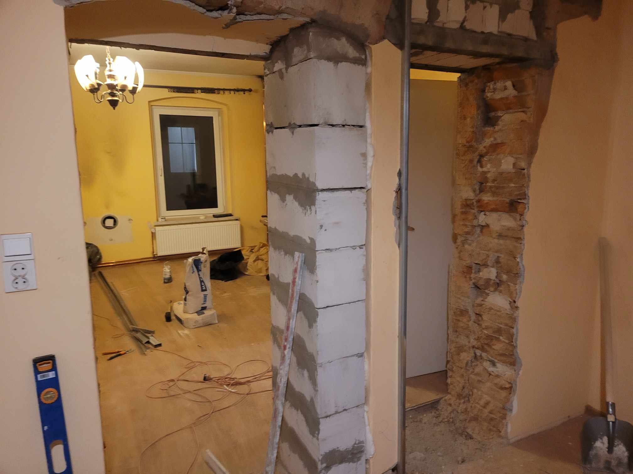 Wnętrze w trakcie remontu: murowana kolumna z betonu komórkowego, odsłonięta cegła, widoczne narzędzia i materiały budowlane na podłodze, żółte ściany w tle.