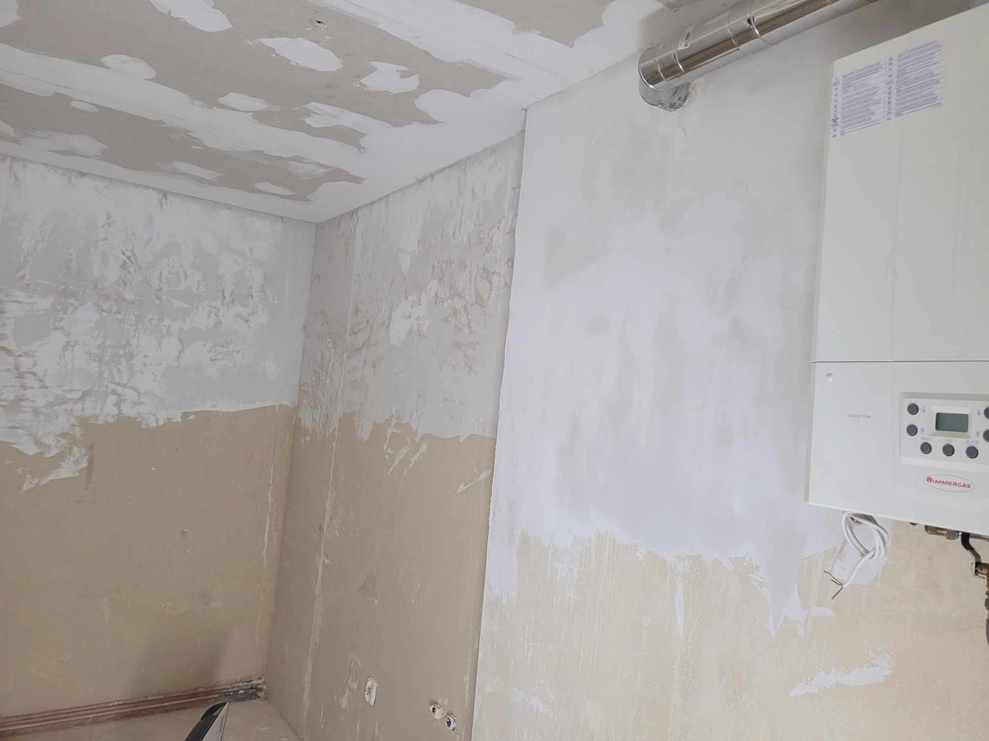 Wnętrze w trakcie remontu: częściowo otynkowane ściany, surowy sufit z widocznymi łatami gipsu, wiszący piec gazowy Immergas z widocznymi przewodami i rurami.
