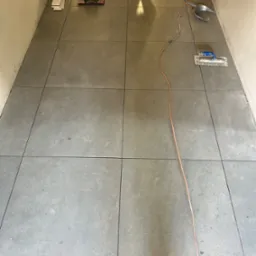 Ułożona podłoga z szarych, kwadratowych płytek, widoczne narzędzia glazurnicze: przecinarka do płytek, paca ząbkowana, szpachelka, kabel elektryczny i odkurzacz przemysłowy.