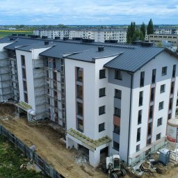 Norbert Siarzynski budownictwo - Wysoki budynek mieszkalny w trakcie budowy, z widocznymi rusztowaniami. Uwagę zwraca zbiornik z zaprawą na placu budowy i częściowo wykończona elewacja w tonacji bieli i szarości.