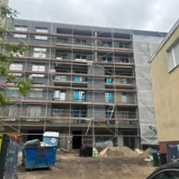 Wielopiętrowy budynek w trakcie prac elewacyjnych. Widoczne rusztowania, częściowo ocieplona ściana, okna zabezpieczone folią. Prace w toku, widoczny pracownik.