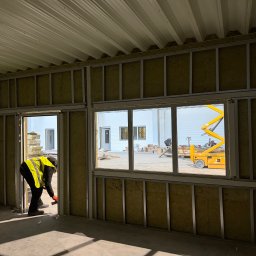 Norbert Siarzynski budownictwo - Montaż izolacji z wełny mineralnej w metalowej konstrukcji szkieletowej. Widoczny pracownik w kamizelce odblaskowej i podnośnik koszowy za oknem.