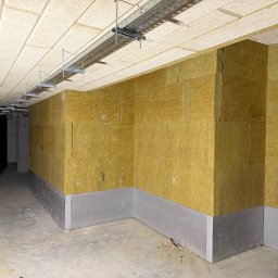Norbert Siarzynski budownictwo - Ocieplone ściany wełną mineralną w surowym wnętrzu. Widoczne szyny pod sufitem i fragment ściany z bloczków. Szare płyty przy podłodze. Etap prac wykończeniowych.