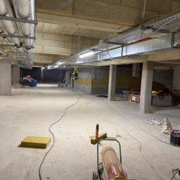 Norbert Siarzynski budownictwo - Wnętrze w trakcie prac wykończeniowych: widoczne ocieplenie stropu wełną mineralną, instalacje pod sufitem, betonowe kolumny i narzędzia budowlane na posadzce.