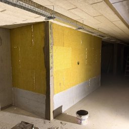 Norbert Siarzynski budownictwo - Ocieplenie ścian wełną mineralną wewnątrz budynku, widoczne profile pod instalacje elektryczne pod sufitem, grunt w wiaderku na podłodze. Etap prac wykończeniowych.
