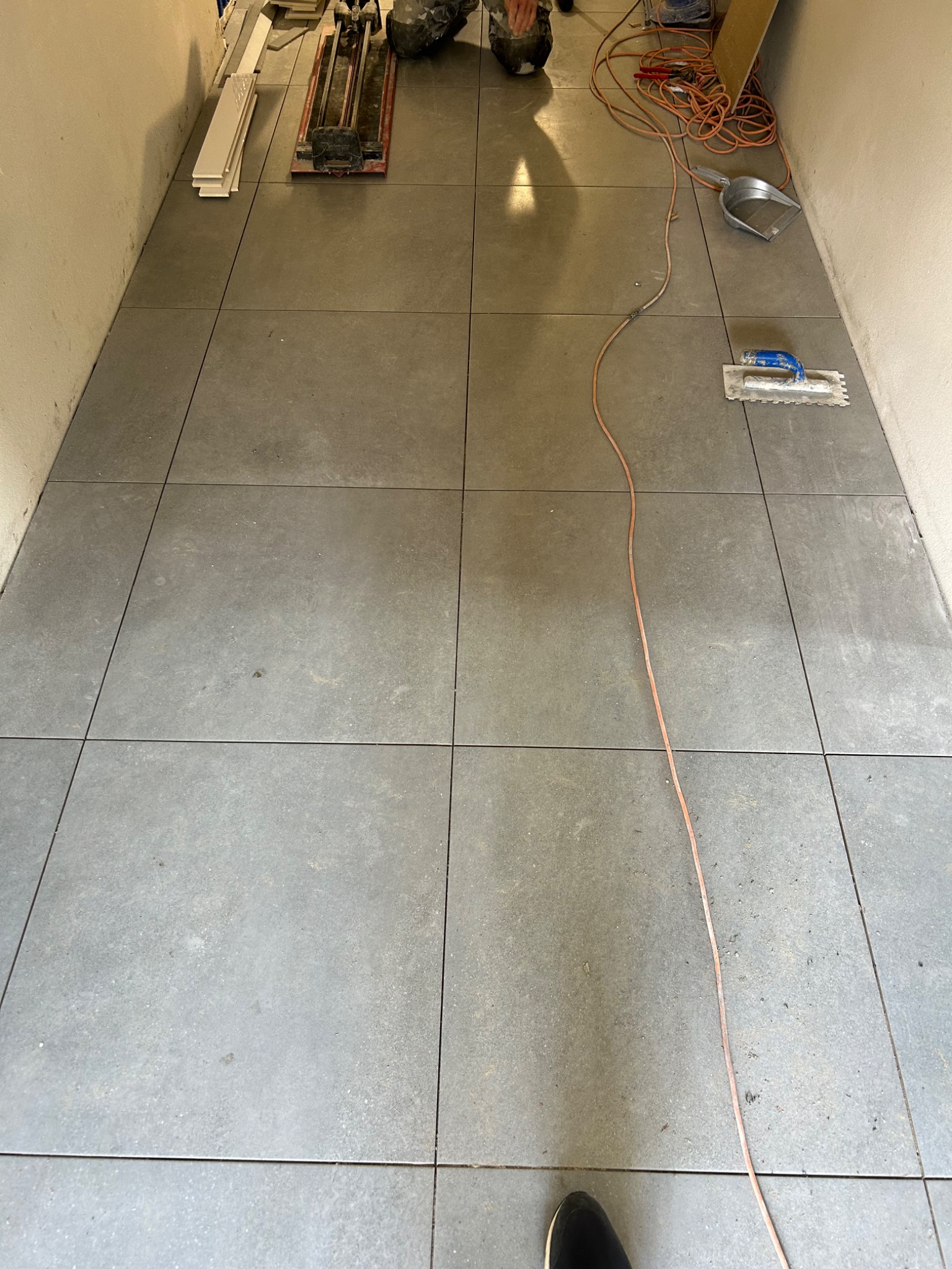 Ułożona podłoga z szarych, kwadratowych płytek, widoczne narzędzia glazurnicze: przecinarka do płytek, paca ząbkowana, szpachelka, kabel elektryczny i odkurzacz przemysłowy.