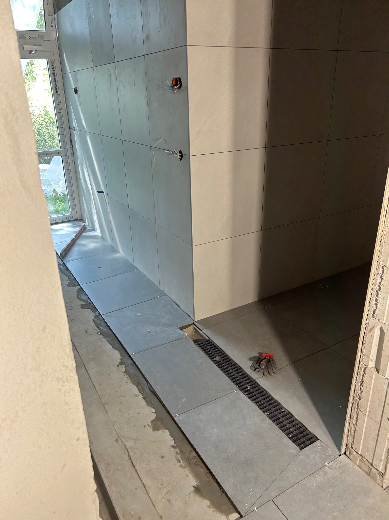 Układanie szarych płytek podłogowych w łazience z widocznym odpływem liniowym i roboczymi rękawicami, w trakcie prac wykończeniowych.